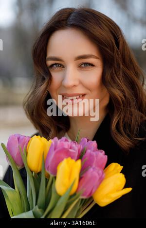 Eine junge Frau mit gewellten braunen Haaren strahlt fröhlich, während sie einen bunten Tulpenstrauß in ihren Armen schmiegt und sich im warmen Glanz eines sonnigen Tages sonnt Stockfoto