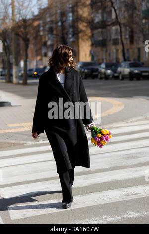 Eine stilvolle Frau geht selbstbewusst über einen Stadtübergang, gebündelt in einem schwarzen Mantel. Ihr heller Tulpenstrauß verleiht dem urbanen lan eine lebendige Note Stockfoto
