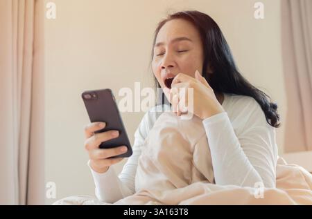Aufnahme einer reiferen Frau, die morgens im Bett das Handy benutzt, während sie gähnt und immer noch schläfrig ist. Selektiver Fokus. Stockfoto