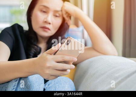 Erwachsene asiatische Frau, die sich langweilig oder traurig fühlt, während sie ihr Smartphone im Wohnzimmer zu Hause spielt. Life-Konzept. Selektiver Fokus. Stockfoto