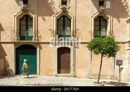 Italien, Sizilien, Ragusa, Stadt, die von der UNESCO zum Weltkulturerbe erklärt wurde, Frau, die am Palazzo (Palast) Bertini vorbeigeht, erbaut im Barock-Stil im 18. Jahrhundert in Ragusa Superiore (hohe Ragusa) auf dem Corso (Straße) Italia, die drei lächerlichen Gesichter an der Fassade sind typisch für den barocken Stil und repräsentieren von links nach rechts den Bettler, den Adligen und den Mann aus dem Osten Stockfoto