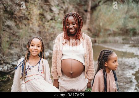 Schwarze schwangere Frau mit Töchtern draußen am Fluss Stockfoto