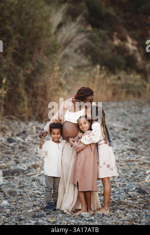 Lächelnde schwarze schwangere Mami-Mix-Rassenkinder küssen Bauch auf Felsen Stockfoto