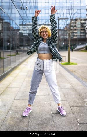 Sorglose junge Frau tanzt mit erhobenen Armen im Stadtzentrum, genießt urbane Musik und drückt Freude aus Stockfoto