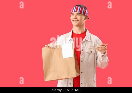 Junger Mann mit Papiertüte mit asiatischem Essen und Stäbchen auf rotem Hintergrund Stockfoto