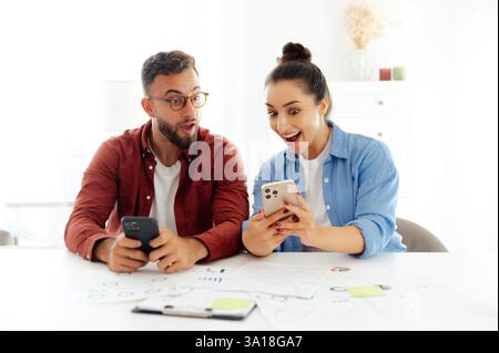 Schockiert überrascht Mann und Frau sitzen am Schreibtisch im Büro, benutzten ihre Smartphones, lasen unerwartete Nachrichten, erhielten atemberaubende Informationen, demonstrierten fröhliche Emotionen und Erstaunen Stockfoto