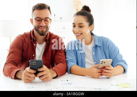 Ein Mann und eine Frau in lässiger Kleidung sitzen an einem Schreibtisch im Büro, nutzen während der Arbeitszeit Smartphones, senden online Nachrichten mit Kunden oder Freunden, surfen E-Mails, lesen Nachrichten, suchen nach Informationen Stockfoto