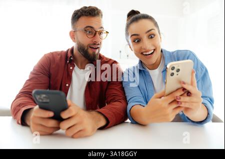 Schockierter Mann und Frau sitzen am Schreibtisch im Büro, benutzen ihre Smartphones, schauen staunend auf Bildschirme, lesen unerwartete Nachrichten, erhielten atemberaubende Informationen, Demonstrationen glückliche Emotionen Stockfoto
