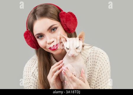 Schöne Frau, die rote flauschige Ohrenschützer trägt und ein haarloses Katzentier umarmt Stockfoto