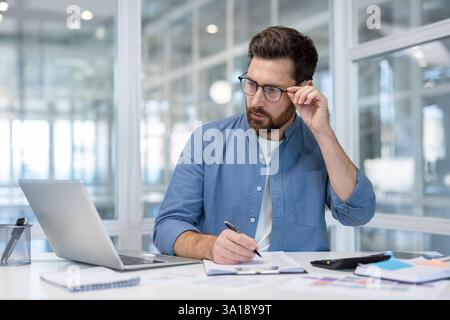 Ernsthaft denkender Buchhalter, der mit Computer im Büro arbeitet. Mann macht Papierkram, füllt Steuerformulare aus. Geschäftsmann, der einen Bericht erstellt, Dokumente im Büro unterschreibt. Stockfoto