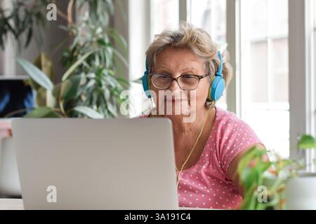 Ältere Frau mit Kopfhörern, die mit einem Laptop am Schreibtisch sitzt Stockfoto