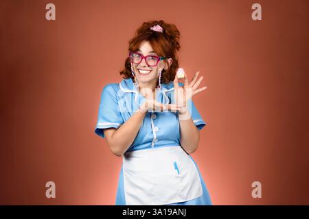 Junge lächelnde rothaarige Kellnerin, die mit rosa Brille, himmelblauem Kleid und weißer Schürze in die Kamera blickte. Einen Cupcake mit einer Hand halten und darauf zeigen Stockfoto