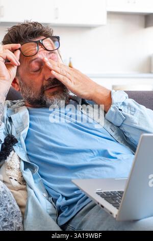 Ein müder Geschäftsmann zieht die Brille ab und spürt die Augenbelastung, wenn er online mit einem Laptop arbeitet, ein Kaukasier sitzt auf dem Sofa und reibt sich die Augen, fühlt Kopfschmerzen und Energielosigkeit Stockfoto