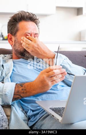 Ein müder Geschäftsmann zieht die Brille ab und spürt die Augenbelastung, wenn er online mit einem Laptop arbeitet, ein Kaukasier sitzt auf dem Sofa und reibt sich die Augen, fühlt Kopfschmerzen und Energielosigkeit Stockfoto