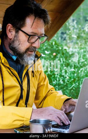 Mann, der gerne draußen mit Laptop arbeitet und in den Bergen des Naturparks sitzt. Konzept der Fernarbeit oder des Lebensstils von Freiberuflern. Freiheit vom Büro. Gutaussehender Mann, der Computer an einem alternativen Arbeitsplatz benutzt Stockfoto