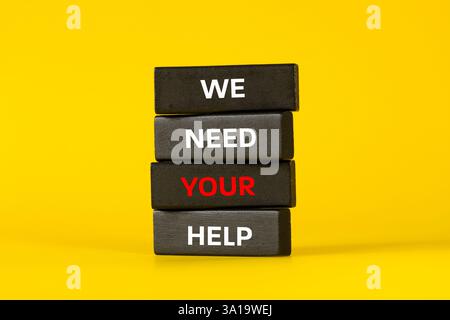 Wir brauchen deine Hilfe, Worte auf schwarzen Holzblöcken geschrieben, gelber Hintergrund. Geschäftliche Nutzung. Wir bitten um Ihre Professionalität und Hilfe Stockfoto