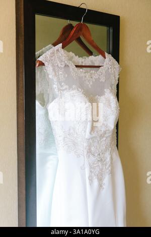 Ein atemberaubendes Hochzeitskleid hängt auf einem hölzernen Kleiderbügel und spiegelt die Spitzendetails im hellen Spiegel des Zimmers wider. Stockfoto