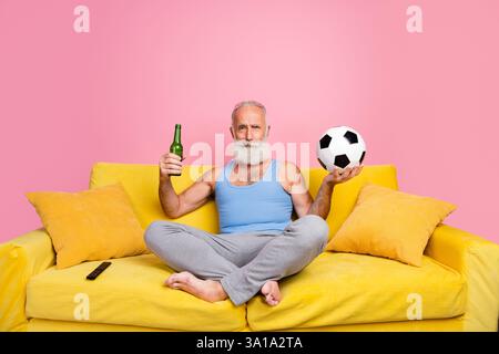 Lächelnder reifer Mann, der sich drinnen auf einem lebhaften gelben Sofa entspannt und einen Fußball und eine Flasche in einer ungezwungenen Umgebung hält Stockfoto