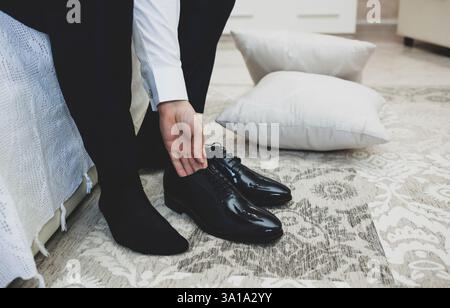 Ein Mann im Anzug bindet die Schnürsenkel auf schwarz klassisch elegante Schuhe. Bräutigam Morgen im Hotel Zimmer vor der Trauung. Stockfoto