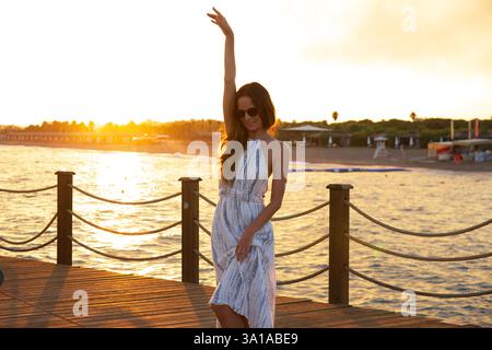 Fröhliche Frau im Sommerkleid posiert am Pier bei Sonnenuntergang am Meer Stockfoto