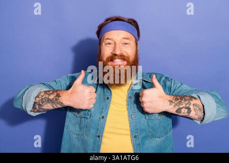 Fröhlicher bärtiger Mann mit Tätowierungen, die die Daumen in lässigem Stil vor violettem Hintergrund hochlegen und Retro- und Hipster-Ästhetik verkörpern. Stockfoto