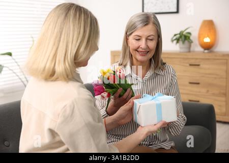 Lächelnde Mutter erhält einen Tulpenstrauß und ein Geschenk von ihrer Tochter auf dem Sofa zu Hause. Alles Gute zum Muttertag Stockfoto