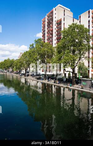 Die Menschen entspannen sich entlang des Canal Saint-Martin in Paris, Frankreich, und genießen den Blick auf moderne Wohnhäuser. Sonniger Tag. Stockfoto