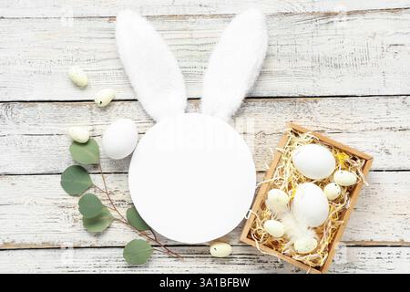 Leere Karte mit Hasenohren, Korb und Ostereiern auf weißem hölzernem Hintergrund Stockfoto