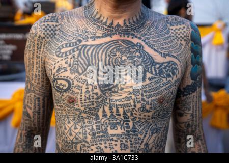 Bang Phra, Nakhon Pathom, Thailand. März 2025. Ein Mann mit Sak Yant Tätowierungen, die seinen Oberkörper bedecken, steht während der jährlichen Wai Kru Zeremonie im Wat Bang Phra in Nakhon Pathom, Thailand. Der Tempel Wat Bang Phra, 40 km von der thailändischen Hauptstadt Bangkok entfernt, findet einmal im Jahr die Wai Gru Zeremonie statt. Der Name des Tempels wird übersetzt mit Kloster des Flussufers Buddha Bild. Sak Yant Tätowierungen sind hochstilisierte Tätowierungen buddhistischer Gebete und religiöser Symbole, die den Gläubigen, die sie auf ihren Körper legen, einen besonderen Schutz und andere Vorteile bieten. Viele Teilnehmer befinden sich in einem Zustand von s Stockfoto