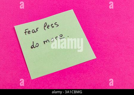 Ein grüner Haftnotiz mit dem Motivationssatz „Fear less do more“ wird auf einem hellrosa Hintergrund platziert. Diese einfache Erinnerung ermutigt zum Handeln Stockfoto