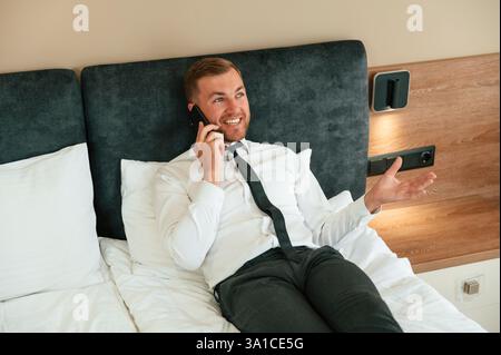 Spaß haben, indem man telefonisch spricht und sich auf das Bett legt. Geschäftsmann ist drinnen im Hotelzimmer. Stockfoto