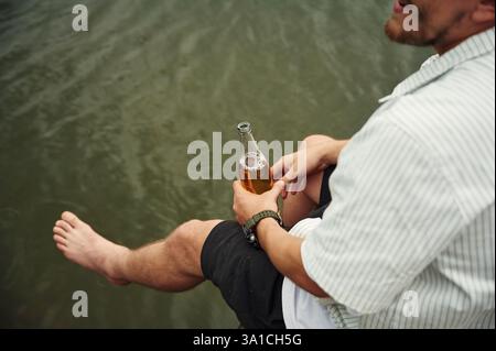 Nahaufnahme eines Mannes, der am Wasser sitzt, mit Bier in der Hand. Stockfoto
