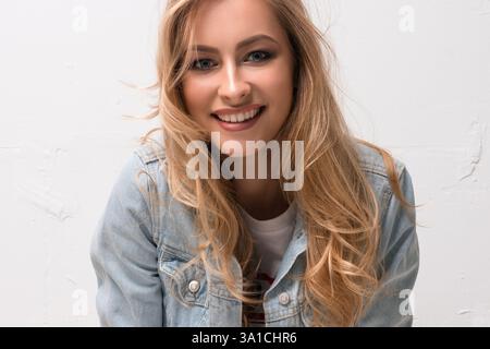 Junge Frau lächelt hell in lässiger Jeansjacke vor hellem Hintergrund Stockfoto