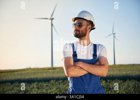 Seriöser Servicetechniker mit Sonnenbrille steht mit gekreuzten Armen auf dem Feld mit Windmühlen. Stockfoto