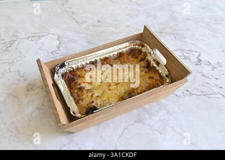 Ein Tablett mit Lebensmitteln steht auf einer Marmorarbeitsfläche. Das Essen ist mit Käse bedeckt und er ist eine Art Lasagne. Das Tablett besteht aus Holz und wird auf einem mar platziert Stockfoto