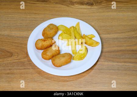 Ein Teller mit gebratenem Huhn und Pommes frites steht auf einem Holztisch. Der Teller ist weiß und das Essen ist goldbraun. Die Pommes frites werden in Keile geschnitten und t Stockfoto