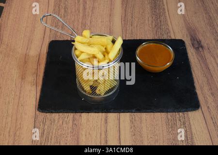 Ein Teller Pommes frites wird mit einer Dippsoße serviert. Die Pommes sind in einem Metallkorb und die Soße in einer Glasschale. Das Bild vermittelt ein beiläufiges A Stockfoto