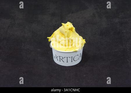 Ein kleiner weißer Becher mit einer Eiskugel darin, darauf steht das Wort „Artica“. Das Eis scheint gelb zu sein und sitzt auf einem schwarzen Su Stockfoto