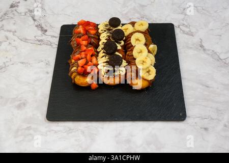 Drei Donuts mit verschiedenen Belägen, einschließlich Bananen und Erdbeeren. Die Donuts sind auf einer schwarzen Platte angeordnet Stockfoto