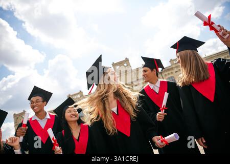 Fröhliche Studenten feiern ihren Abschluss in schwarzen Gewändern und roten Akzenten an einem sonnigen Tag Stockfoto