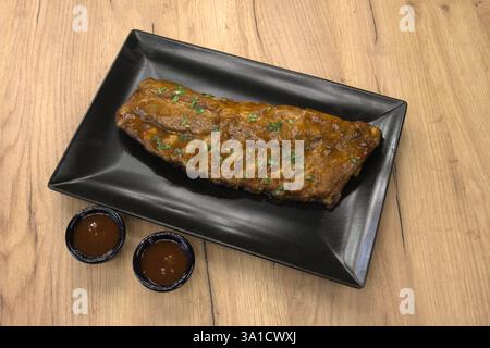 Ein großes Stück Fleisch liegt auf einem schwarzen Teller mit einer Sauce. Das Fleisch ist gekocht und er ist Rippchen. Der Teller wird auf einem Holztisch aufgestellt Stockfoto