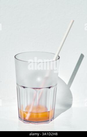 Minimalistisches Glas mit gestreiftem Stroh und orangen Getränkeresten, auf einer weißen Oberfläche in sauberer und heller Beleuchtung. Stockfoto