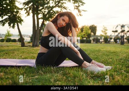 Auf der Yogamatte gegen Sonnenlicht sitzen. Schöne junge Frau in sportlicher Kleidung ist tagsüber im Park. Stockfoto
