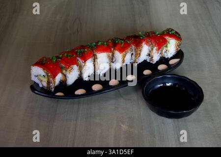 Eine lange rote Sushi-Rolle sitzt auf einem schwarzen Teller neben einer kleinen schwarzen Schüssel mit Soße. Die Sushi-Brötchen sind mit roter Sauce bedeckt und haben verschiedene Beläge Stockfoto
