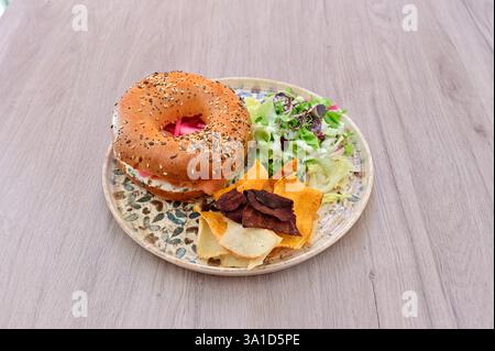 Ein Teller mit Essen mit einem Sandwich, Pommes und Salat. Das Sandwich ist ein Bagel mit einem Aufstrich. Der Salat ist eine Mischung aus Gemüse und Gemüse. Die Platte Stockfoto