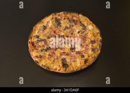 Eine Pizza mit Schinken und Käse drauf. Die Pizza steht auf einem schwarzen Tisch. Die Pizza ist sehr groß und hat viele Beläge Stockfoto