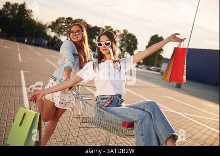 Fahren in einem Einkaufswagen. Zwei schöne Frauen in lässiger Kleidung halten draußen Taschen. Stockfoto