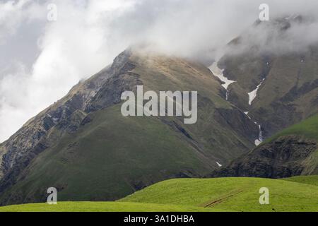 Majestätische Berglandschaft mit üppig grünen Hängen und zerklüfteten felsigen Gipfeln, die teilweise von Nebelwolken bedeckt sind. Ein kleiner Wasserfall stürzt das valle hinunter Stockfoto