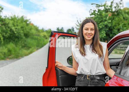 Eine lächelnde Brünette in einem weißen Hemd und einer schwarzen Jeans steht neben einem roten Auto Stockfoto