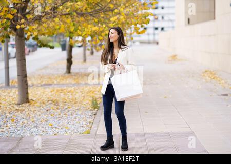 Eine junge Frau, die gemütlich eine schöne Herbststraße entlang schlendert und eine Einkaufstasche hält Stockfoto